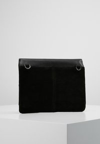 Černá semišová a kožená taška plochého tvaru, s akcenty ze stříbrného kování a dvěma kruhovými kroužky na přední straně pro upevnění.