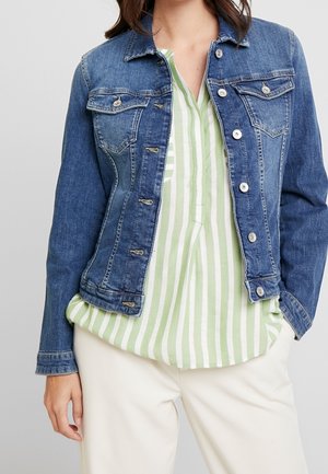 Vrouw die een blauwe spijkerjas draagt over een groen-wit verticale gestreepte blouse en witte broek, staand met één ontspannen hand.