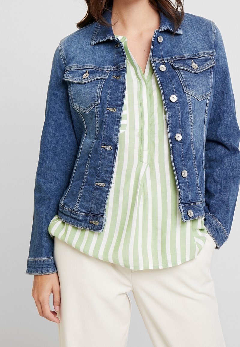 Femme portant une veste en jean bleue par-dessus une chemise rayée verticale verte et blanche et un pantalon blanc, debout avec une main détendue.
