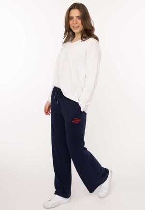 Frau mit weißem Sweatshirt und marineblauer Jogginghose mit „Feel Good Club“-Logo, steht mit einem leicht angehobenen Bein und lächelt.