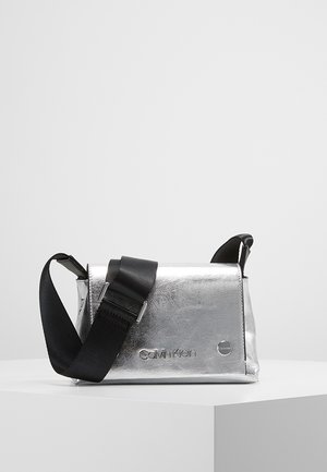 Sac bandoulière métallique argenté Calvin Klein avec large sangle noire réglable sur un socle blanc devant un fond gris.