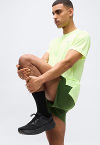 Άνδρας με νέον κίτρινο πουκάμισο, πράσινα σορτς και μαύρα αθλητικά παπούτσια τεντώνει το δεξί γόνατο προς το στήθος πάνω σε απλό λευκό φόντο.