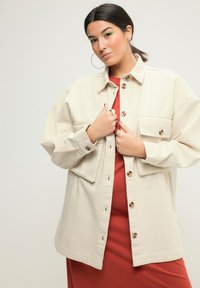 Beige oversized shirtjacket met twee voorkruiszakken en bruin knoopaccenten, gelaagd over een rode jurk met een gladde textuur.
