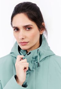 Ljus teal zip-jacka med huva, med justerbara snören, en slät textur och en subtil logotyp på sidan.