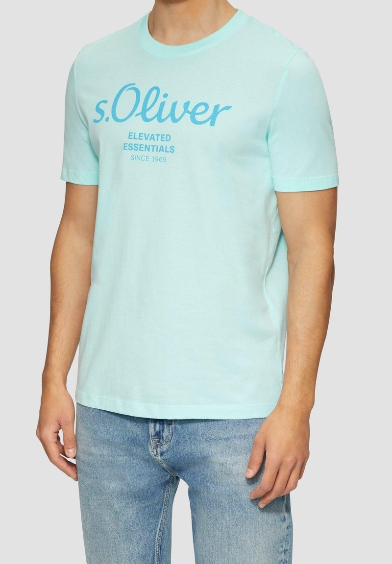 Homme portant un T-shirt s.Oliver bleu clair avec le texte "Elevated Essentials Since 1969" et un jean bleu, debout devant un fond uni.