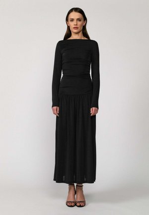 Kobieta stojąca na jednolitym tle, ubrana w długą, czarną dopasowaną sukienkę z długim rękawem oraz czarne sandały na wysokim obcasie z paskami.