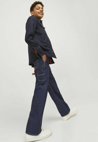 Temno moder jeans jakna z rahlo sproščenim krojem, z črnimi gumbi in stranskimi razporki. Kombinirana z širokimi jeans hlačami in belimi supergami.