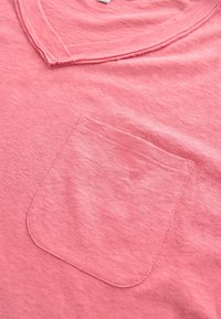Rosa Baumwoll-T-Shirt mit V-Ausschnitt und einer einzelnen Brusttasche mit sichtbaren Nähten. Der Stoff hat eine weiche, strukturierte Oberfläche.