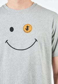 T-shirt in cotone grigio con una grande grafica di un viso sorridente nero e una patch circolare arancione "Save the Duck" sul petto.