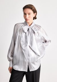 Silvery, satin-like blouse with a high neckline, bow tie detail, and billowy sleeves. Smooth texture, fluid design, and flowing silhouette.