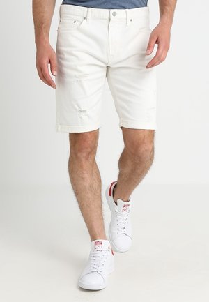 Man wearing white rolled-hem denim shorts and white sneakers walking on a plain white background.