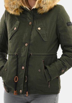 Femme portant une parka d'hiver vert olive avec col en fausse fourrure, boutons sur le devant, taille à cordon, poches zippées et écusson en tissu sur la manche.