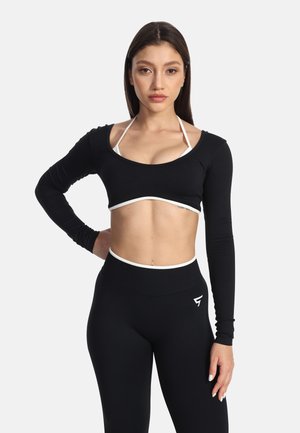 Mladá žena má na sebe čierny crop top s dlhým rukávom cez bielu športovú podprsenku a čierne legíny s vysokým pásom, stojí s jednou rukou na boku.
