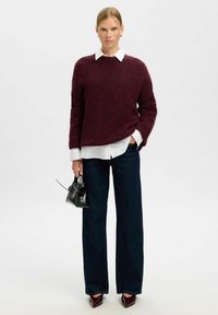Bordeauxfarbener Wollpullover mit Rundhalsausschnitt, über ein weißes Hemd mit Kragen getragen. Hochgeschnittene dunkelblaue Jeans und schwarze Handtasche mit Akzenten.