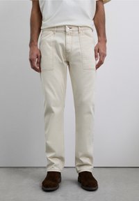 Hombre vestido con pantalones rectos color blanco roto, con bolsillos de parche y zapatos de ante marrones, de pie ante un fondo neutro.