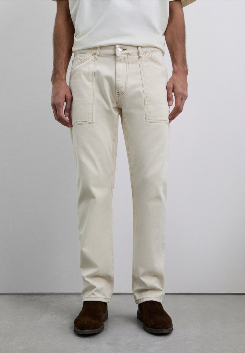 Hombre vestido con pantalones rectos color blanco roto, con bolsillos de parche y zapatos de ante marrones, de pie ante un fondo neutro.