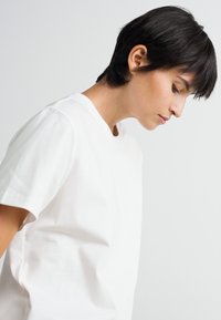 Ung person med kort mørkt hår iført en simpel hvid T-shirt, der ser nedad mod en ensfarvet lys grå baggrund.