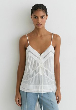 Vrouw met gevlochten haar die een witte mouwloze blouse met kantdetails en lichtblauwe spijkerbroek draagt, staand tegen een effen witte muur.