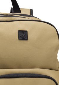 Mochila beige hecha de tela duradera con detalles en negro en la cremallera, que cuenta con un parche de logo texturizado y correas ajustables.