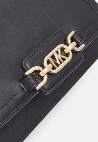 Dettaglio di una borsa in pelle nera con catena dorata, caratterizzata da un logo MK intrecciato su un'etichetta rettangolare cucita.