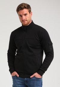 Giacca nera con zip e colletto alto, presenta una cucitura orizzontale sul petto e una tasca sul braccio sinistro. Tessuto strutturato.