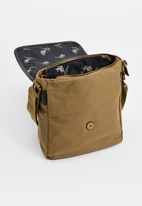 Ruskea kankaasta valmistettu olkalaukku, jossa on vetoketjullinen suljenta, musta sisävuori, jossa toistuva kuvio, ja etupuolella nappikoriste.