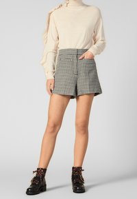 Beige pitkähihaisessa paidassa on ribbikuvio ja röyhelöyksityiskohtia, se on yhdistetty musta-valkoruutuisiin shortseihin ja mustiin nauhakenkiin nilkkamalleissa.