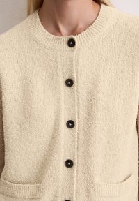 Kremowy kardigan o fakturze przypominającej śmietanę, z okrągłym dekoltem, pięcioma czarnymi guzikami i dwiema przednimi kieszeniami. Miękka tkanina o lekko puszystym wyglądzie.