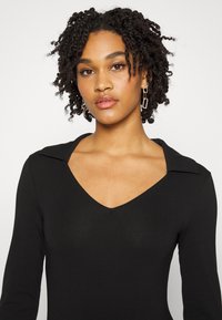 Vestido preto de mangas longas com decote em V e gola. Tecido suave com design ajustado. Modelo usa brincos retangulares em destaque.