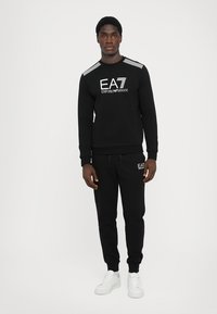 Black sweatshirt and matching joggers made of soft fabric, featuring white logo and striped accents, complemented by white sneakers.