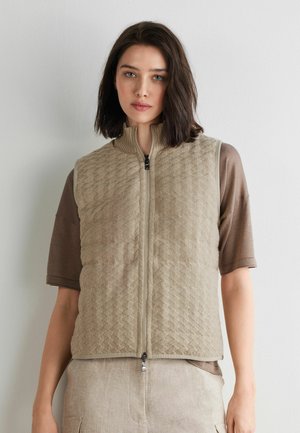 Vrouw met donker haar tot op de schouders, gekleed in een beige gewatteerde bodywarmer met rits, over een bruin T-shirt met korte mouwen en lichtbeige broek.