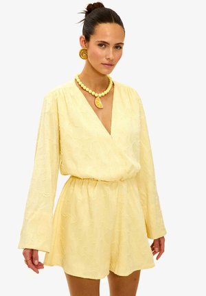 Femme aux cheveux en chignon portant une combishort texturée jaune pâle, de grandes boucles d'oreilles spirales en or, et un collier de perles jaune avec un pendentif en forme de tranche d'orange.