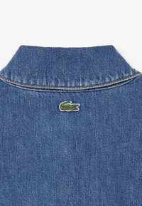 Col de veste en denim bleu avec un petit logo de crocodile vert brodé sous la nuque au dos.