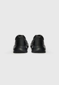 Chaussures de sport noires avec une surface lisse, un talon arrondi et une semelle texturée. Design minimaliste avec des coutures renforcées au niveau du talon.