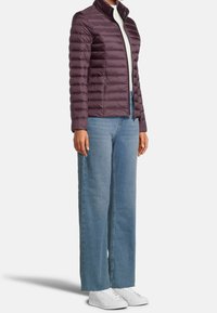 Vycpaná bunda švestkové barvy s stojacím límcem a horizontálním prošitím, kombinovaná s světle modrými džínami s širokými nohavicemi a bílými teniskami.