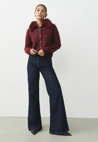 Bordeaux fuzzy cardigan met een bontkraag, vijf knopen, geribbelde manchetten, in combinatie met donkere spijkerflared jeans en schoenen met een spitse neus.