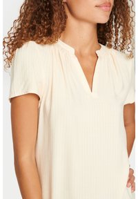 Ljusbeige kortärmad topp med V-ringning, strukturerad tyg med ett subtilt vågmönster. Rynkade detaljer vid halsringningen.