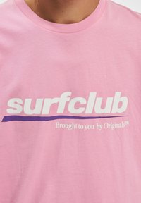 Rosa bomulls T-skjorte med fet hvit tekst "surfclub" og lilla linje under, med mindre tekst "Brought to you by Originals™."