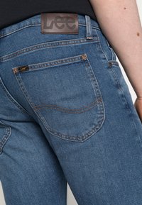 Fato de ganga azul com uma etiqueta de couro castanho em relevo, costura laranja em contraste e um único bolso traseiro com uma etiqueta.