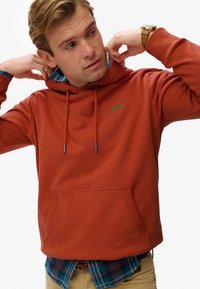 Rust-colored hoodie with a front pocket, drawstring hood, and embroidered logo. Layered over a blue plaid shirt, featuring ribbed cuffs.