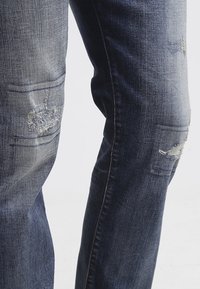 Zbliżenie na zużyte niebieskie dżinsy z postrzępionymi łatami na udach i kolanach na jednolitym tle.