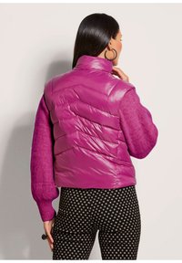 Lila Puffer-Weste mit einem Chevron-Steppmuster, hohem Kragen und kontrastierenden langen, flauschigen lila Ärmeln. Gemusterte schwarze Hosen mit weißen Quadraten.