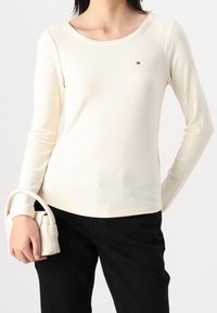 Camiseta de manga larga de color crema con canalé, escote amplio y pequeño detalle de logo, combinada con pantalones negros y un bolso mini.