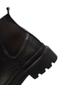 Botín de cuero negro con panel de malla texturizada en negro, suela de goma gruesa y un patrón de tracción pronunciado. Presenta un corto elástico lateral para facilitar su uso.