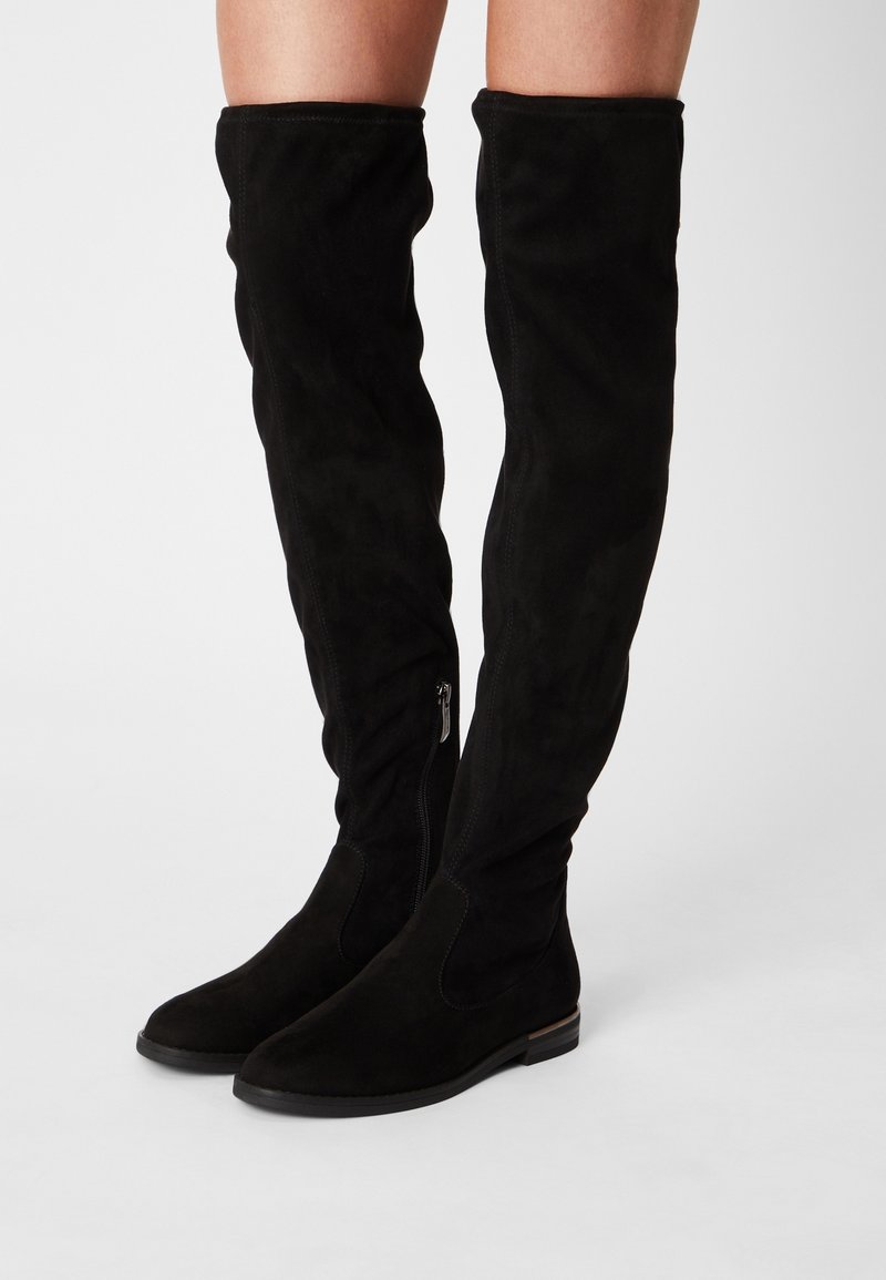 Bottes noires en suede au-dessus du genou avec un bout affûté, un talon plat et un zip latéral pour un enfilage facile. Texture lisse avec une silhouette épurée.