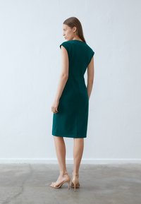 Topsložen aqua haljina s kratkim rukavima, dužine do koljena, s glatkom teksturom i minimalnim detaljima. Model nosi bež sandale s visokom petom.