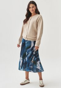 Beige gebreide cardigan met een ronde halslijn en knopen aan de voorkant, gecombineerd met een blauwe geplooide rok met abstracte patronen en witte sneakers.