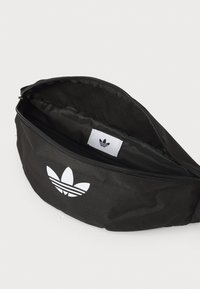 Fekete szövet deréktáska fehér Adidas logóval és három csíkkal. A cipzáras záródás egy bélelt belső teret tár fel, amelyen márkajelzés található. Kompakt, kerekdizájn.