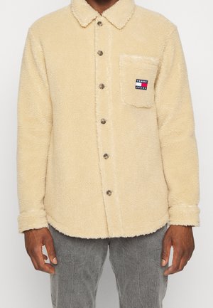 Beige Fleecejacke zum Knöpfen mit Kragen und einer Brusttasche mit Tommy Jeans-Logo, getragen zu grauen Hosen.