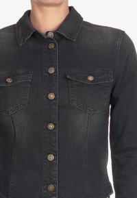 Svart denimjacka med knapp-up framsida, två bröstfickor och mässingsknappar. Har en klassisk krage och texturerad tyg.